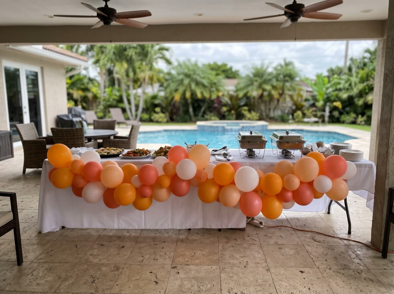 Organic balloon garland in mango, coral and white along a buffet on a covered Miami pool patio